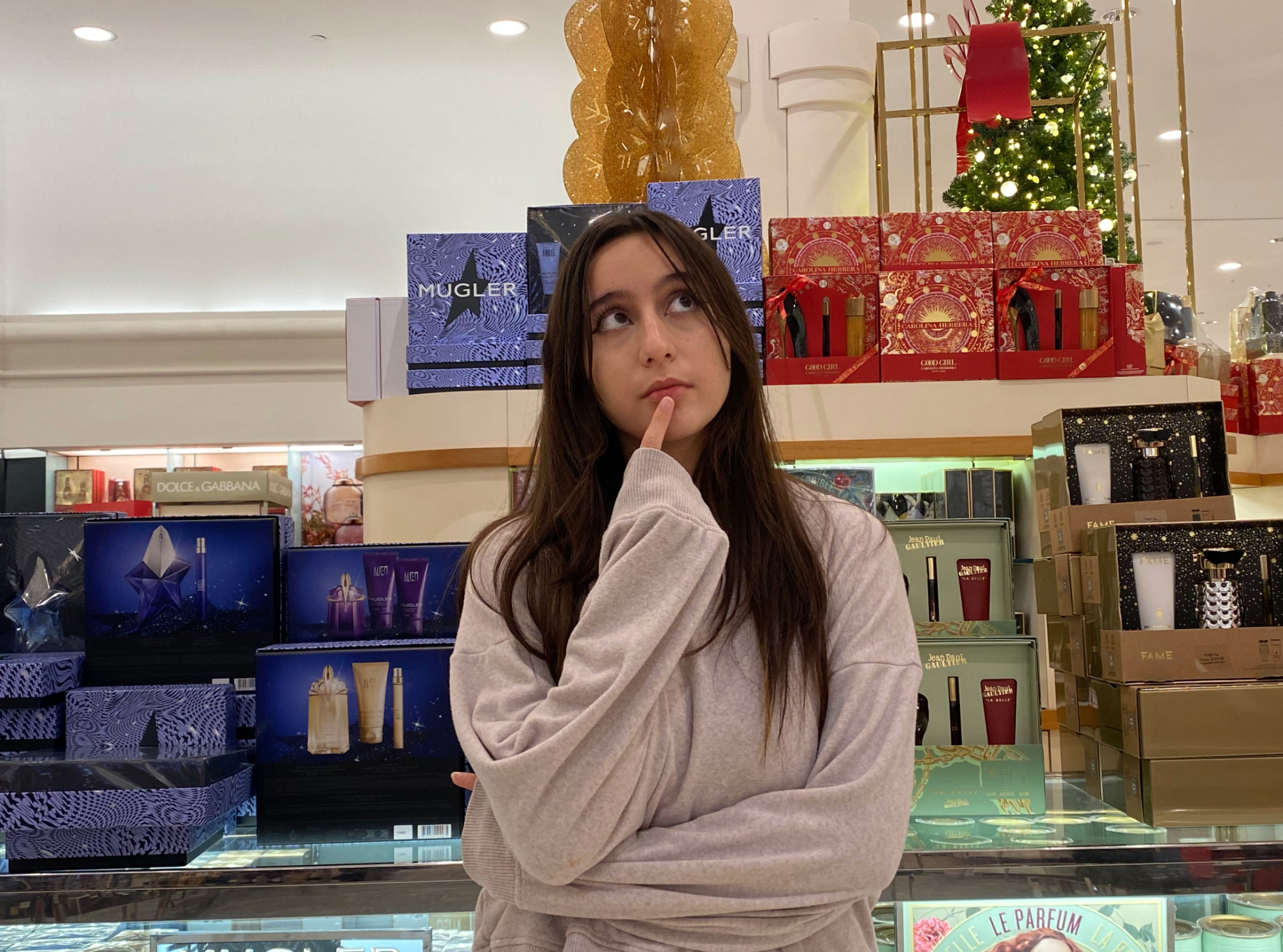 Ceylin Golcu stands in front of a gift set display stand, wondering which one to pick. The display includes high-end brands like Mugler, with the perfume costing around $100.