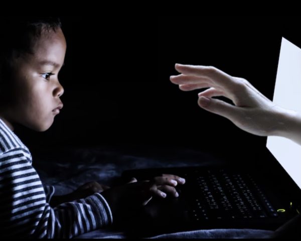 A little kid playing on his computer while a hand reaches out to him from a white screen.